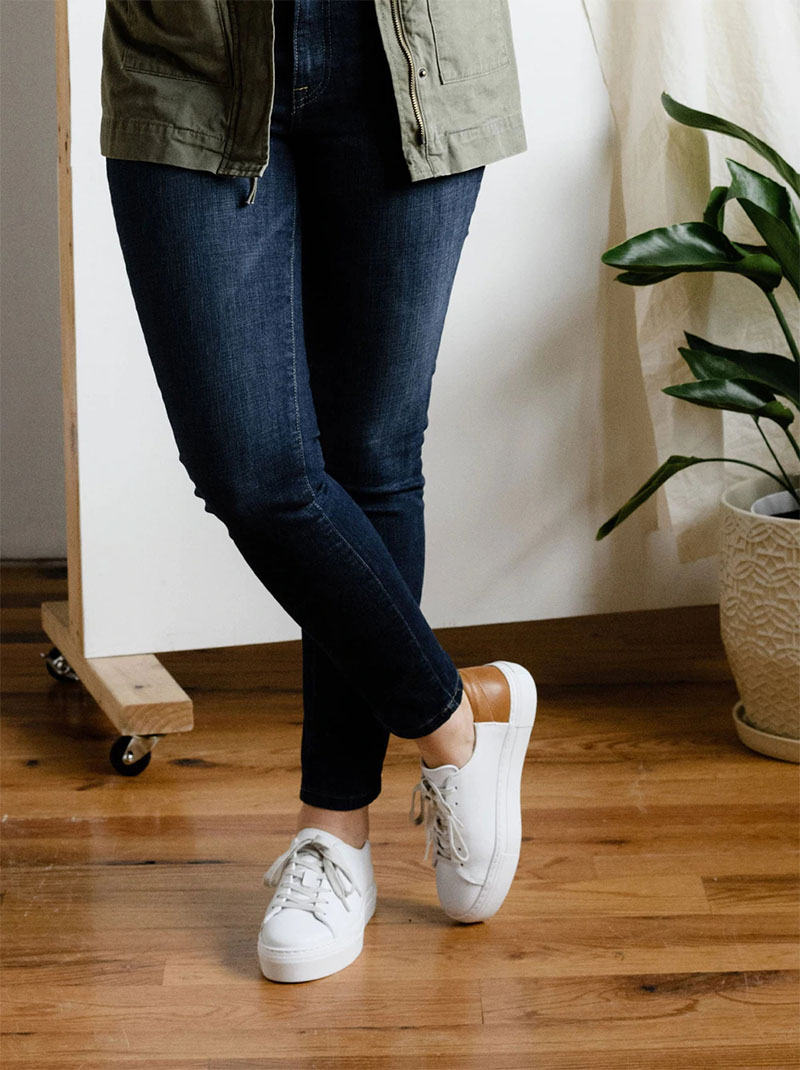 Woman wearing ABLE Emmy Sneakers and blue jeans standing on a wooden floor near a potted plant